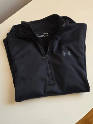 Svart halvzip från Under Armour - Säljer en svart tröja från Under Armour med halv dragkedja framtill och diskret logga på bröstet. Tillverkad i ett mjukt och stretchigt funktionsmaterial som passar perfekt för träning och sportaktiviteter.