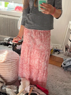 Rosa långkjol med paisleymönster - Säljer en flowy rosa långkjol med vitt paisleymönster. Kjolen har en avslappnad passform och resår i midjan för extra komfort. Perfekt för dig som gillar bohemisk stil och vill ha något luftigt till sommaren. Den är lågmidjade i storlek 158, men ganska lång. Från Gina