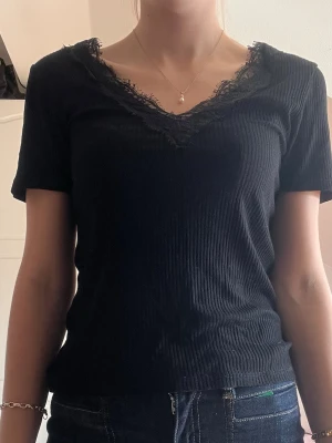Svart ribbad topp med spetsdetalj - Svart ribbad topp med v-ringning och spetsdetaljer både fram och bak. Kortärmad och figurnära passform, perfekt till jeans eller kjol. Toppens material känns mjukt och stretchigt.