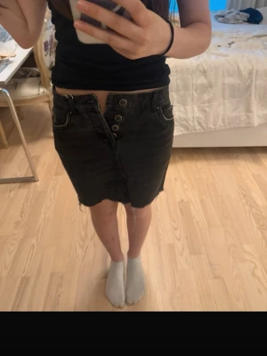 Svart jeanskjol från ONLY - Säljer en svart jeanskjol från ONLY i modell 'Raw Life Reg'. Kjolen har hög midja, flera knappar framtill och rå kant nertill för en edgy look. Tillverkad i ekologisk bomull och har klassiska fickor både fram och bak.