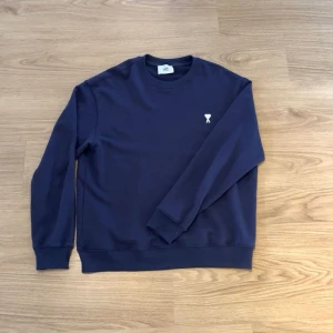 Mörkblå sweatshirt med hjärtdetalj - Stilren mörkblå sweatshirt med rund hals och ribbade muddar. På bröstet finns en liten vit broderad detalj med ett hjärta och bokstaven A. Perfekt till jeans eller över en skjorta för en avslappnad look.