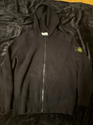 Svart stickad hoodie från Stone Island - Säljer en svart stickad hoodie från Stone Island med dragkedja framtill och ikonisk patch på vänster ärm. Tröjan har ribbad struktur och huva, perfekt för dig som gillar streetwear och vill ha något både snyggt och bekvämt.