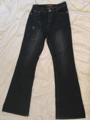 Mörkblå bootcut jeans med pärlor - Säljer ett par mörkblå bootcut jeans med dekorativa pärlor och broderier på vänster lår. Jeansen har klassisk femficksmodell och stängs med knapp och dragkedja. Materialet är denim i bomull med lätt stretch.