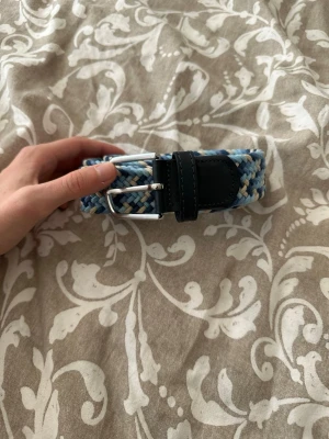 Flätat blått bälte från ONEWear - Snyggt flätat bälte från ONEWear i olika blå nyanser, beige och vitt. Bältet har detaljer i svart skinn och ett silvrigt spänne. Perfekt för att ge din outfit en cool touch och passar till både jeans och chinos. Bältet är 100cm 