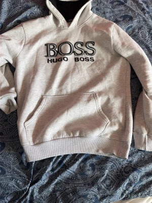 Hugo Boss hoodie - Snygg grå hoodie från Hugo Boss med stor logga broderad på bröstet. Hoodien har en klassisk känguruficka framtill, ribbade muddar och en mjuk insida. Huvan är fodrad i svart tyg vilket ger en schysst kontrast. Perfekt för en avslappnad och trendig look.