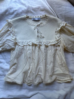 Krämvit romantisk blus från ZARA - Söt krämvit blus från ZARA med virkade detaljer upptill och volangkant. Blusen har knytband vid halsen och puffiga långärmar. Materialet är tunt och luftigt, perfekt för en bohemisk look. Passar dig som gillar vintageinspirerat mode. Liten i storlek💞