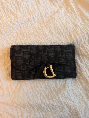 Svart monogramplånbok med gulddetalj - Snygg svart plånbok med monogrammönster och en stor guldfärgad D-detalj på framsidan. Plånboken har en rektangulär form och stängs med lock. Materialet ser ut att vara textil med tryckt mönster.