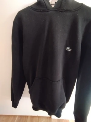 Lacoste hoodie  - Snygg svart hoodie från Lacoste med klassisk krokodillogga på bröstet. Hoodien har huva och en stor magficka framtill. Tillverkad i mjukt material som känns skönt mot huden. Perfekt för en avslappnad och stilren look.