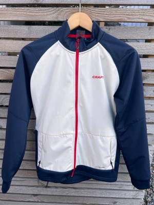 Blåvit träningsjacka från Craft - Core Warm XC Jacket Jr är en funktionell jacka med vind- och vattenavvisande front och borstad trikå i rygg och sidor designad för längdskidåkning och liknande aktiviteter i kalla förhållanden.  Denna mångsidiga träningsjacka har ett återvunnet 2-lagers vind- och vattenavvisandet material fram och på ärmarna som ger bra väderskydd samt en funktionell återvunnen trikå i rygg och sidor för effektiv ventilation och reglering av kroppstemperaturen. 