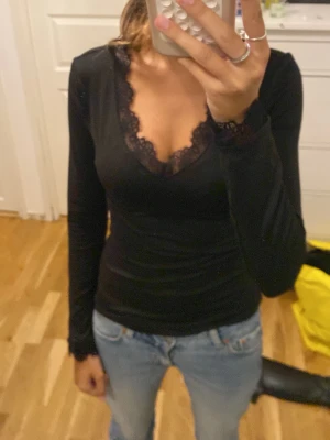 Svart långärmad topp med spets - Snygg svart långärmad topp med v-ringning och spetsdetaljer vid halsringning och ärmslut. Figurnära passform som framhäver siluetten. Perfekt att styla med jeans eller kjol för en trendig look.