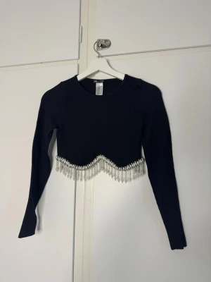 Svart crop top med pärldetaljer - Trendig svart långärmad crop top med rund halsringning och dekorativa pärlor längs nederkanten. Ribbad struktur i tyget ger en snygg look och pärlfransen gör tröjan extra unik. Perfekt för dig som vill sticka ut lite extra.