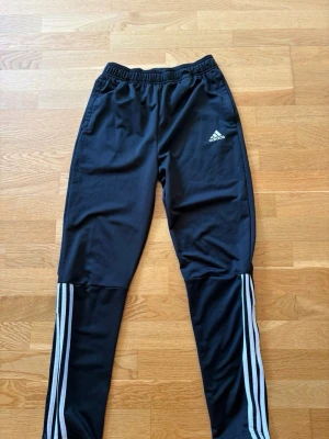 Svarta Adidas träningsbyxor - Säljer ett par svarta träningsbyxor från Adidas med klassiska vita ränder längs benen och Adidas-logga på låret. Byxorna har resår i midjan och dragkedjor vid bensluten. Perfekta för träning eller chill.