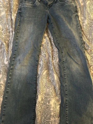Blå bootcut jeans valerie - Snygga blå bootcut jeans med lätt slitna detaljer och klassisk femficksmodell. Jeansen har en något utsvängd passform nedtill och är tillverkade i ett mjukt denimtyg. Perfekta för en avslappnad och trendig look.