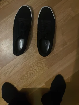 Svarta låga sneakers med vit sula - Stilrena svarta sneakers med klassisk vit sula och snörning. Skorna har en enkel och clean design som passar till det mesta. Materialet ser ut att vara textil eller mockaimitation. Perfekta för dig som gillar minimalistisk stil.
