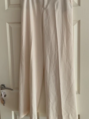Vida beige kostymbyxor från Zara - Snygga vida kostymbyxor från Zara i beige med matchande bälte och rund spänne. Byxorna har hög midja och är tillverkade i ett mjukt, vävt material som ger en stilren look. Perfekta för dig som gillar en clean och modern stil.