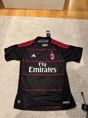 AC Milan retro tröja #11 Ibrahimović - Snygg svart AC Milan matchtröja från Adidas med röda detaljer och klubbmärke på bröstet. Tryck med 'Fly Emirates' på framsidan och 'Ibrahimović 11' på ryggen. Tillverkad i lätt och ventilerande material, perfekt för fotboll eller träning.