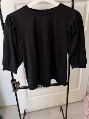 Svart topp med puffärm - En svart topp med puffiga trekvartsärmar och rund halsringning. Tillverkad i mjukt bomullsmaterial som känns skönt mot huden. Enkel men trendig design som passar till många olika stilar.