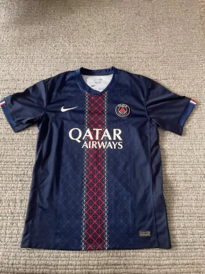 PSG fotbollströja hemmaställ 24/25  - Fräsch PSG fotbollströja från säsongen 24/25. Tröjan är mörkblå med en snygg röd linje i mitten. Man kan enkelt matcha tröjan med andra kläder. Den är tillverkad i lätt och ventilerande material så att den är perfekt till träning.