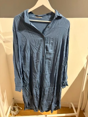 Blå långärmad skjortklänning Sézane - Blå långärmad skjortklänning från Sézane med knappar framtill och klassisk krage. Klänningen har en avslappnad passform och är tillverkad i ett mjukt tyg som ger en skön känsla. Perfekt för dig som gillar enkel och stilren design.