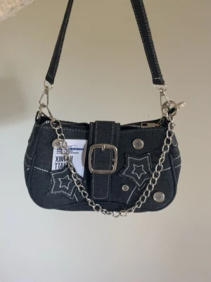 Svart denim handväska med kedjedetaljer - Cool svart handväska i denim med vita sömmar, stjärnpatchar och silverfärgade knappar. Väskan har en bred rem, dekorativ kedja och ett spänne framtill. Invändigt finns ett fack med dragkedja. Perfekt accessoar för en trendig look.