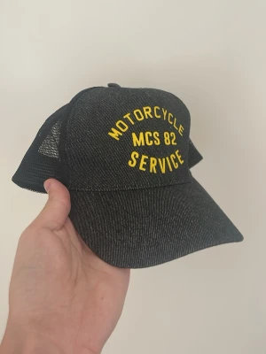 Svart truckerkeps med gul text - Snygg svart truckerkeps med gul brodyr där det står 'MOTORCYCLE MCS 82 SERVICE' på framsidan. Kepsen har böjd skärm och mesh på sidorna och bak för extra ventilation. Justerbar snapback baktill för perfekt passform.