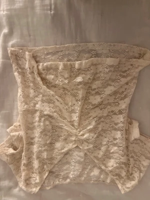 Beige spets topp - Säljer en beige axelbandslös tubtopp i spets med v-ringad detalj framtill. Toppen har ett romantiskt och feminint uttryck med genombrutet spetsmönster och är perfekt för att styla lager på lager.