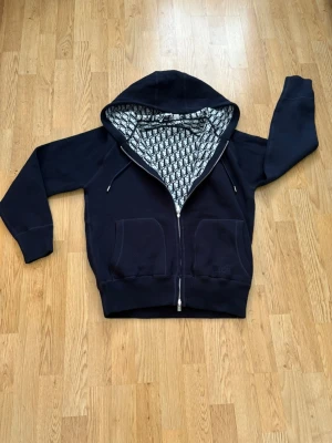 Marinblå d1or ziphoddie - Snygg marinblå hoodie från D1or med dragkedja och två stora fickor framtill. Insidan av huvan har ett ikoniskt svartvitt D1or-mönster som ger en exklusiv vibe. Tillverkad i mjukt material och har justerbara snören i huvan. Perfekt för dig som vill sticka ut.