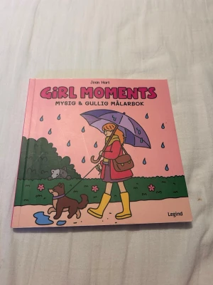 Girl Moments mysig målarbok - En söt och färgglad målarbok med titeln 'Girl Moments' av Jean Hart. Omslaget är rosa med en tecknad tjej i regnkläder och gula stövlar som går med en hund under ett paraply. Perfekt för dig som gillar att färglägga gulliga motiv.