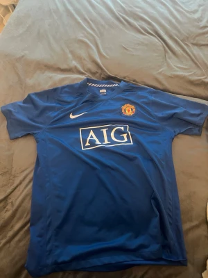 Blå Manchester United tröja Nike - Säljer en blå Manchester United tröja från Nike med AIG-logga på bröstet och klubbmärke på vänster sida. Tröjan har korta ärmar och rund hals, tillverkad i ett lätt och ventilerande material som passar perfekt till träning eller match.