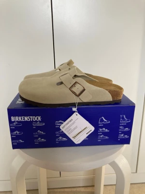 Beige Birkenstock Boston tofflor - Snygga beige Birkenstock Boston sandaler i mocka med justerbart spänne i metall och klassisk korksula. Perfekta slip-ins med mjuk fotbädd för maximal komfort. Passar dig som gillar stilrena och tidlösa skor.