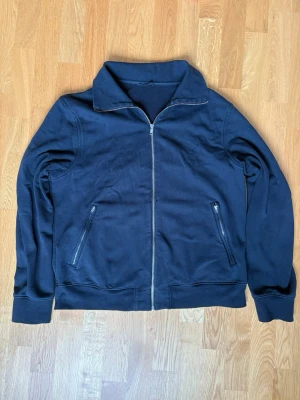 Mörkblå ziptröja med hög krage från Filippa K - En stilren mörkblå ziptröja med hög krage och två fickor med dragkedja framtill. Tröjan kommer från Filippa K och är i storlek L. Tröjan är i ett använt skick. Perfekt för lager-på-lager och enkel att matcha med olika outfits.