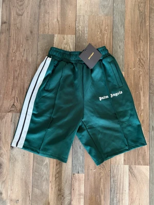 Gröna shorts från Palm Angels - Snygga gröna shorts från Palm Angels med vita sidoränder och logga på vänster ben. Shortsen har elastisk midja och sidofickor. Perfekta för en avslappnad och sportig stil.