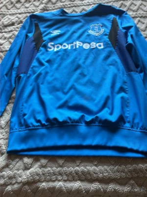 Blå Everton träningsjacka Umbro - Säljer en blå långärmad träningsjacka från Everton FC med Umbro-logga och SportPesa-tryck på bröstet. Jackan har svarta detaljer på sidorna och är tillverkad i ett lätt, sportigt material som passar perfekt till fotbollsträning.