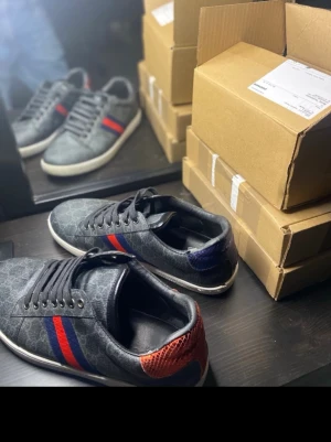 Gucci Ace  - Snygga grå sneakers med GG-mönster, röd och blå rand på sidan samt kontrastdetalj i rött på hälen. Skorna har vita sulor och svarta skosnören. Materialet är en mix av textil och läderliknande yta. Perfekta för dig som vill sticka ut med stilrena detaljer.