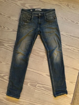 Replay Anbass Aged 5 Years - Snygga blå slim fit jeans med slitna detaljer och ljusa partier på låren och knäna. Klassisk femficksmodell med dragkedja och knapp i midjan. Jeansen har en smal passform och är tillverkade i denim med lätt stretch för extra komfort. W27 L30 men passar W28 L30