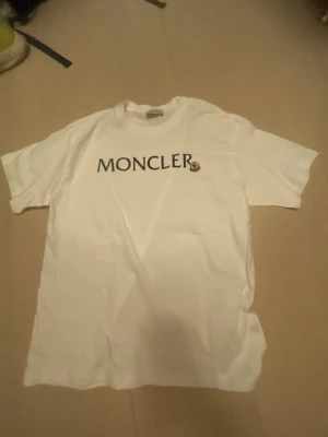 Vit Moncler t-shirt med logga - Säljer en klassisk vit t-shirt från Moncler med stor svart logga tryckt på bröstet och det ikoniska Moncler-emblemet. T-shirten har rund hals och korta ärmar, perfekt till jeans eller shorts.