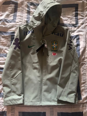 Ljusgrön Brasilien vindjacka med huva - Snygg ljusgrön vindjacka med huva och dragkedja framtill. Jackan har Brasilien-logga, Nike-logga och flera tryck på bröst och ärmar. Perfekt för dig som gillar fotboll och streetstyle. Jackan har två fickor och justerbar huva.