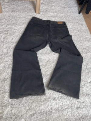 Svarta bootcut jean - Säljer ett par gråa jeans med breda ben och råa, avklippta benslut. Jeansen har klassisk femficksmodell och är tillverkade i denim. Perfekta för en avslappnad och trendig look. OBS hål mellan benen, se bild!