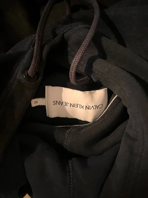 Svart hoodie från Calvin Klein Jeans - Snygg svart hoodie från Calvin Klein Jeans med klassisk huva och dragsko. På bröstet finns en broderad CK-logga i vitt. Hoodien har en stor magficka och är tillverkad i mjukt material, perfekt för en chill och stilren look.
