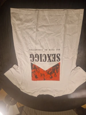 Vit t-shirt med SEXCIGG tryck - Vit t-shirt med svart och rött grafiskt tryck på bröstet där det står SEXCIGG och en spegelvänd text. Klassisk passform och rund hals. Perfekt för dig som gillar statement-plagg och vill sticka ut lite extra.