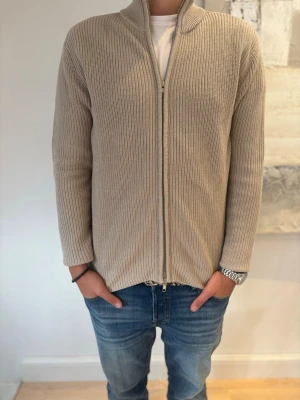 Beige ribbstickad ziptröja - Säljer en beige ribbstickad tröja med dragkedja framtill och hög krage. Tröjan har lång ärm och är gjord i ett mjukt, tjockare material som passar perfekt för kyligare dagar. Enkel och stilren design som är lätt att matcha.