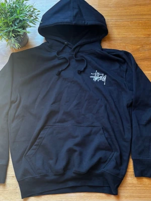 Svart Stüssy hoodie - Svart hoodie från Stüssy med vit logga tryckt på bröstet och ryggen. Klassisk modell med huva, dragsko och stor magficka. Perfekt för en streetwear-look.
