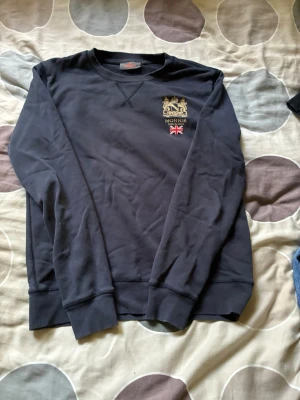 Mörkblå sweatshirt från Morris - Snygg mörkblå sweatshirt från Morris med broderad logga och brittisk flagga på bröstet. Tröjan har rund halsringning, ribbade muddar vid ärmslut och nederkant samt en klassisk college-look. Perfekt för en avslappnad och stilren outfit.