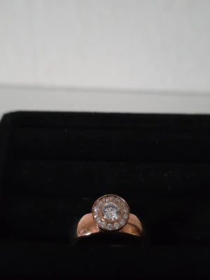 Ingnell Jewellery ring roséguld stål 19 mm - 🌸 Elegant ring från Ingnell Jewellery Sweden i roséfärgat stål. Stilren modell med en större klar sten i mitten, omgiven av mindre glittrande stenar. Fin både till vardags och fest.  🔖 Storlek: 19 mm 🧵 Material: Stål 🎨 Färg: Rosé/roséguld med klara stenar  ✨ Helt ny och oanvänd.  🎁 Vid leverans förpackas ringen fint i en liten presentpåse. Perfekt även som present.