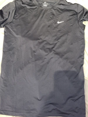 Nike Tshirt - Grå t-shirt från Nike med diskret logga på bröstet. Tillverkad i ett lätt och ventilerande funktionsmaterial, perfekt för träning. Klassisk passform med rund hals och korta ärmar.