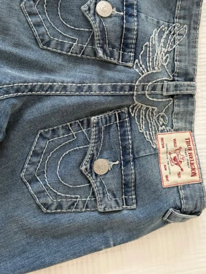 Blå bootcut jeans från True Religion - Säljer ett par blå bootcut jeans från True Religion med tydliga kontrastsömmar och klassisk femficksdesign. Jeansen har en snygg tvättad look och breda benslut som ger en retrovibe. Perfekta för dig som vill sticka ut med en unik jeansstil. Använda en gång och inga tecken på slitage 