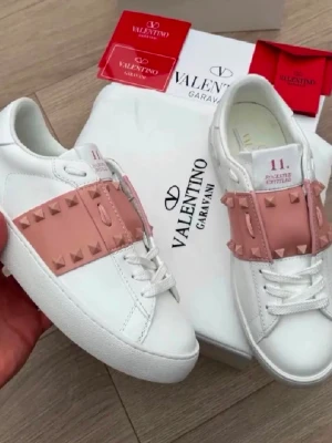 Valentino Garavani vita sneakers - Snygga vita sneakers från Valentino Garavani med breda ljusrosa band dekorerade med nitar över vristen. Skorna har en chunky vit sula med mönstrad undersida och klassisk låg silhuett. Tillverkade i slätt läder för en exklusiv känsla.