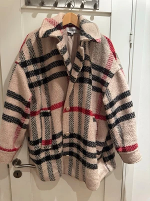 Rutig teddyjacka från Weekday - Säljer en beige teddyjacka från Weekday med rutigt mönster i svart och rött. Jackan har stora knappar framtill, bred krage och två fickor. Materialet är fluffigt och mjukt, perfekt för kalla dagar. Oversized passform för en avslappnad look.