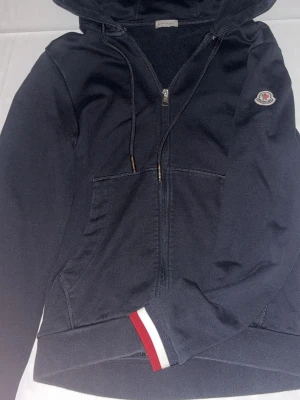 Marinblå zip-hoodie med Moncler-logga - Snygg marinblå hoodie från Moncler med dragkedja, fickor framtill och huva med dragsko. Monclers klassiska logga på bröstet och rödvit detalj på ärmslut och huva. Perfekt för en avslappnad och stilren look.