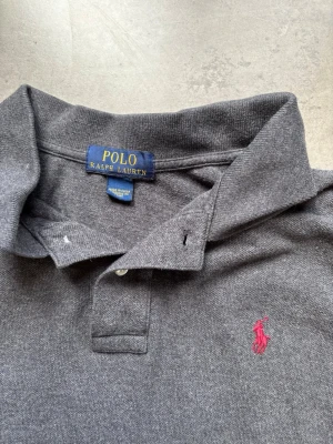Grå långärmad piké från Polo Ralph Lauren - Klassisk grå långärmad pikétröja från Polo Ralph Lauren med krage, knäppning vid halsen och den ikoniska rosa logobrodyr på bröstet. Tillverkad i mjukt bomullsmaterial som känns skönt mot huden. Perfekt för en avslappnad men ändå stilren look.